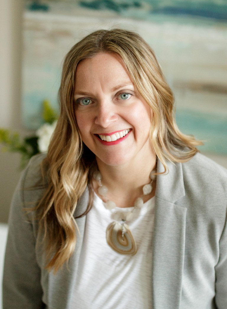 An interior designer with long blond hair and wearing a gray blazer is standing in a room she has designed. She is smiling at the camera.