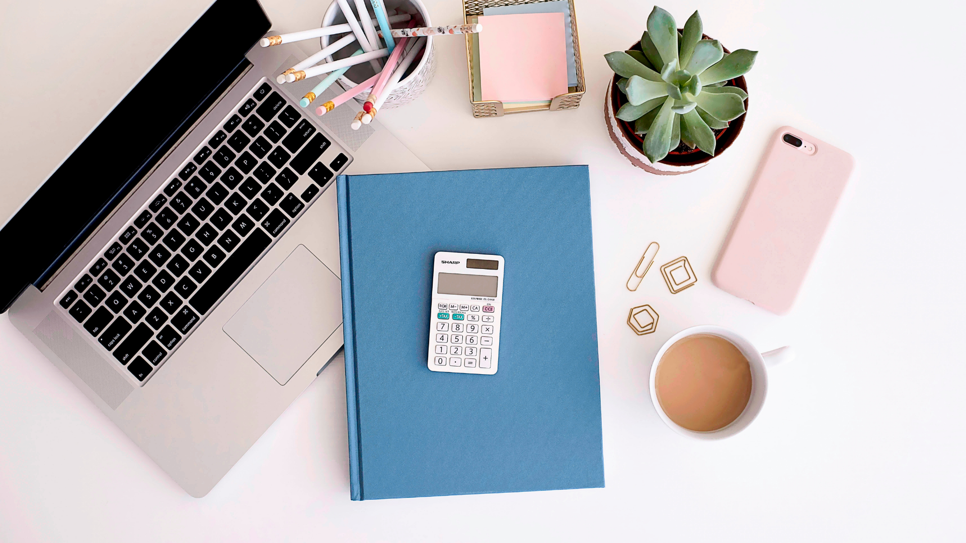 An interior designers desk. A blue notebook sits in the middle of the desk with a pink phone and coffee cup to the right and a computer to the left. There is also a small potted potted plant next to the pink phone.