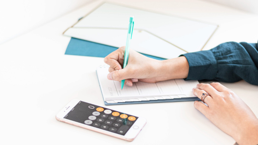 An interior designer in a blue shirt uses a notebook and calculator to plan her interior design discovery call. She is holding a turquoise pen in her right hand.