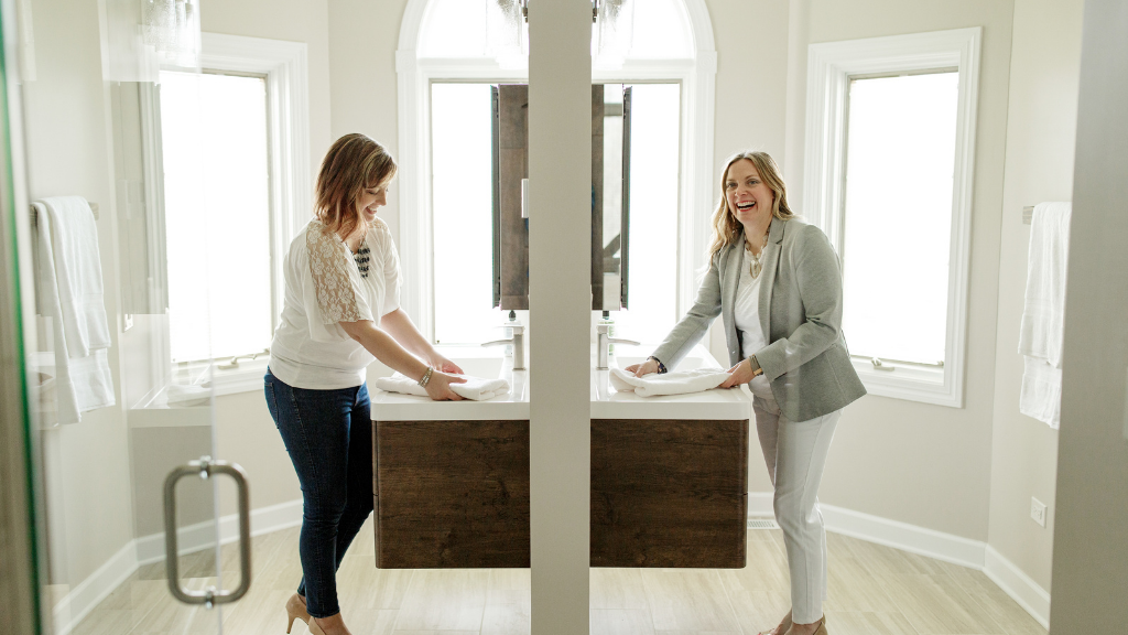 An interior designer reveals her design to the client. They are standing in a bright, airy bathroom. The client is standing at a sink on the left and the interior designer is standing at a sink on the right.