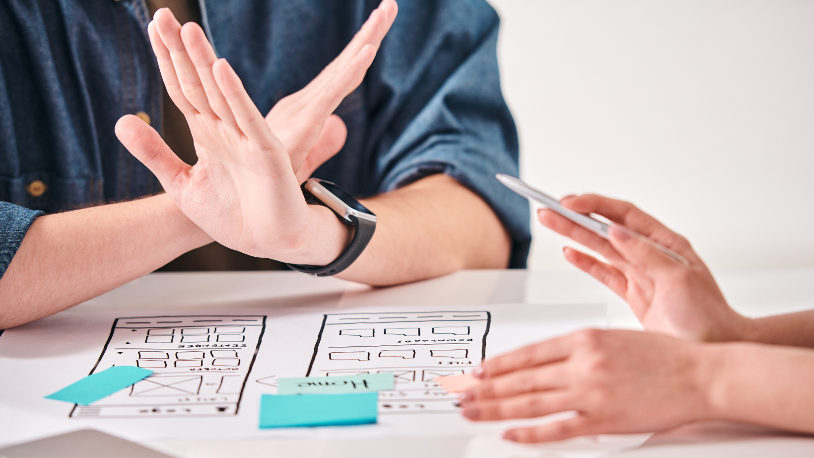 An interior designer in a blue shirt is sitting at a table with her client. her hands are crossed in front of her, as if to signal that she is saying no. There is a blueprint on the table with blue post-it notes.