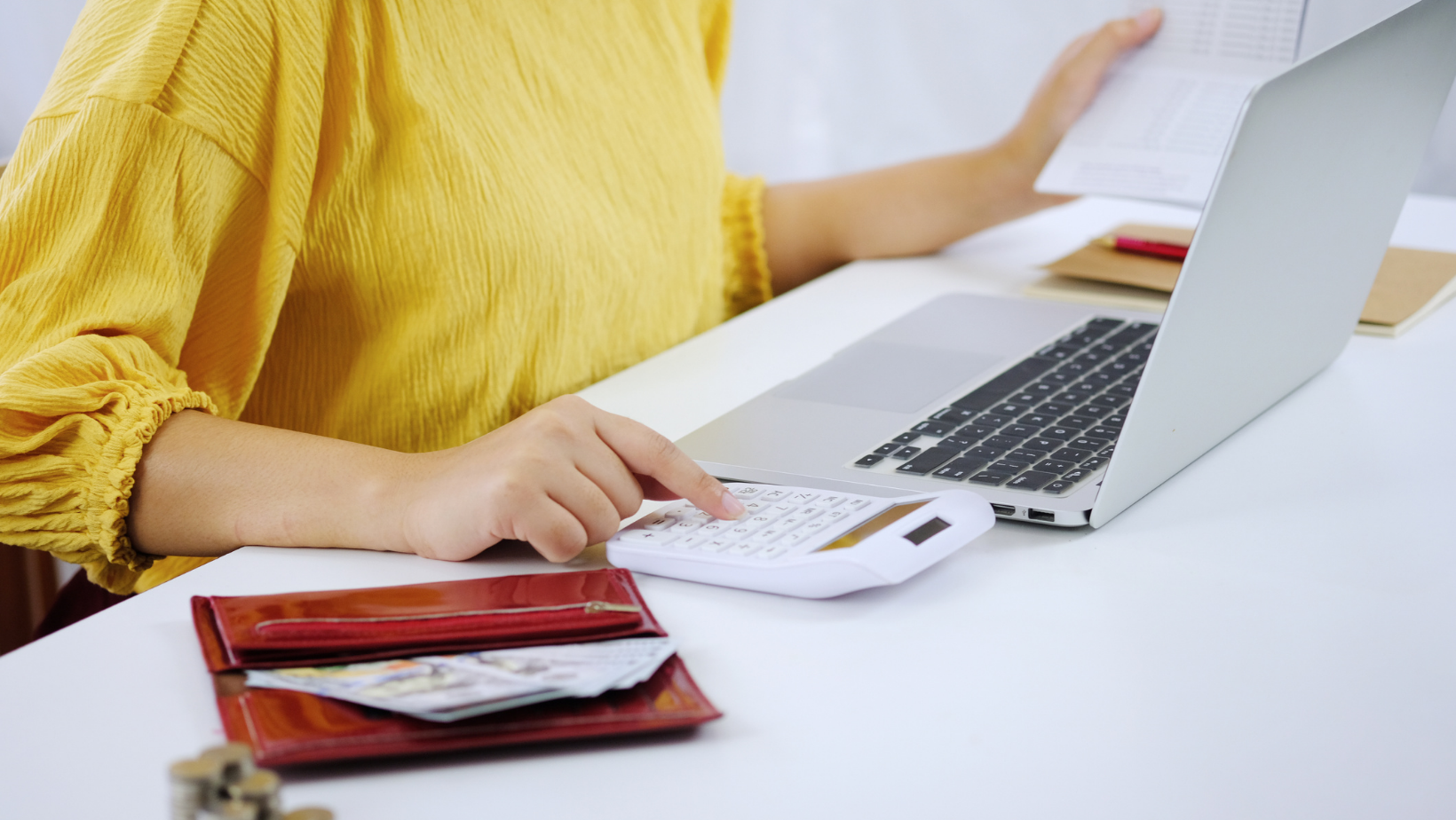 A woman in a bright yellow top is using a calculator and computer to figure out how to raise client prices. Her wallet is open on the table next to her.