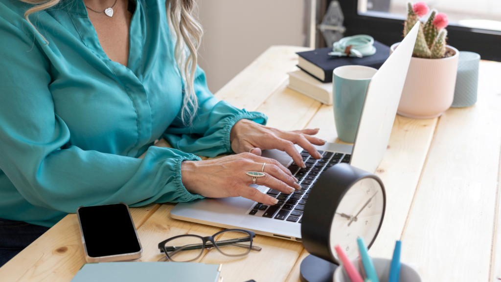 A woman in a bright turquoise blouse is working on her computer, developing vendor relationships. There is a black clock in front of the computer.